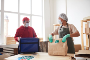homem de boné vermelho e uma bolsa de delivery azul ao lado de uma mulher com touca de cabelo com um embalagens de entrega. ambos estão de máscara de proteção em um pequeno ou medio restaurantes