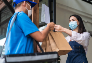 homem vestindo roupa e bone azul entregando uma encomenda de comida para uma mulher sorrindo e morena